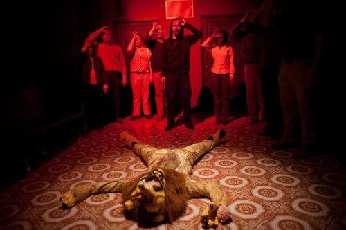 A figure in a lion costume lies spread-eagled on a patterned floor as a crowd salutes in red light