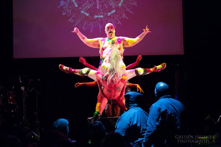 A shirtless man in red face paint and suspenders spreads his arms above a pile of performers in tutus and knee pads