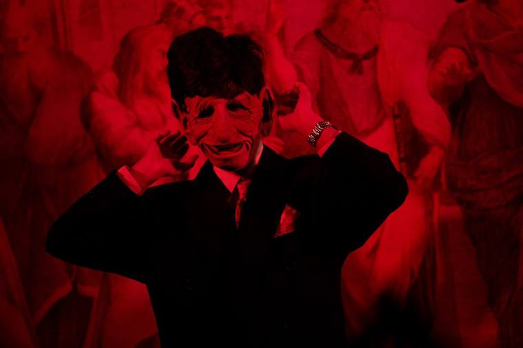 A performer in a grinning rubber mask and suit, bathed in red light
