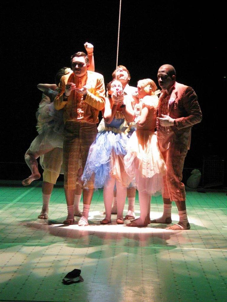 Dancing vs. The Rat Experiment: the cast huddles in a spotlight on a green floor, one dancer in a blue tutu