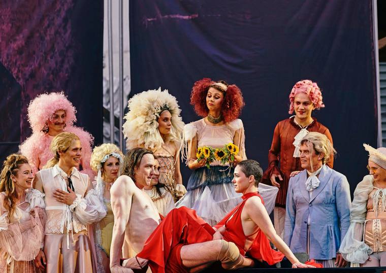 Jedermann at the Salzburg Festival: a crowd in ruffles, wigs and sunflowers gathers around a shirtless man and a woman in red, who lounge in front