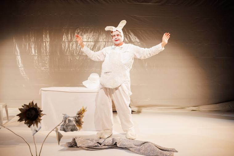 Tony Torn as Spider Rabbit, arms flung wide in a white bunny suit on a white plastic-sheeted stage