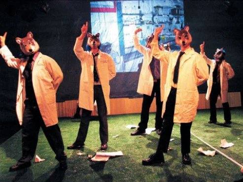 Performers in rat masks and lab coats raise their hands on a green floor