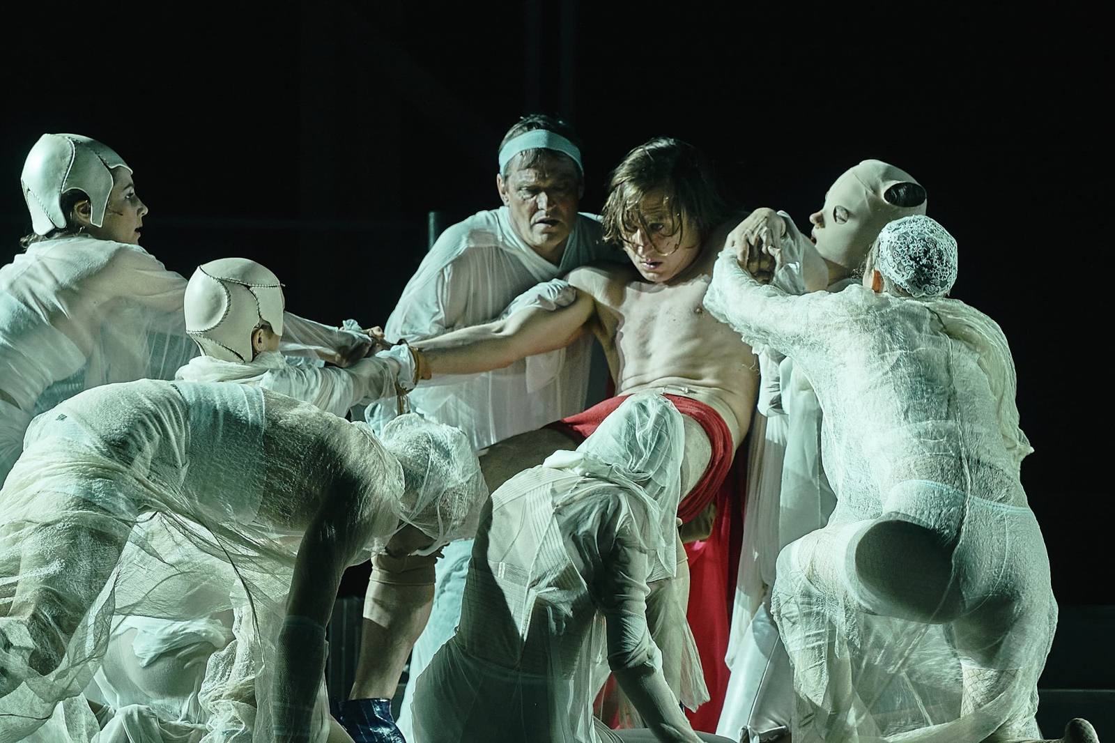 Jedermann at the Salzburg Festival: a shirtless young man in red is held by figures wrapped in white gauze