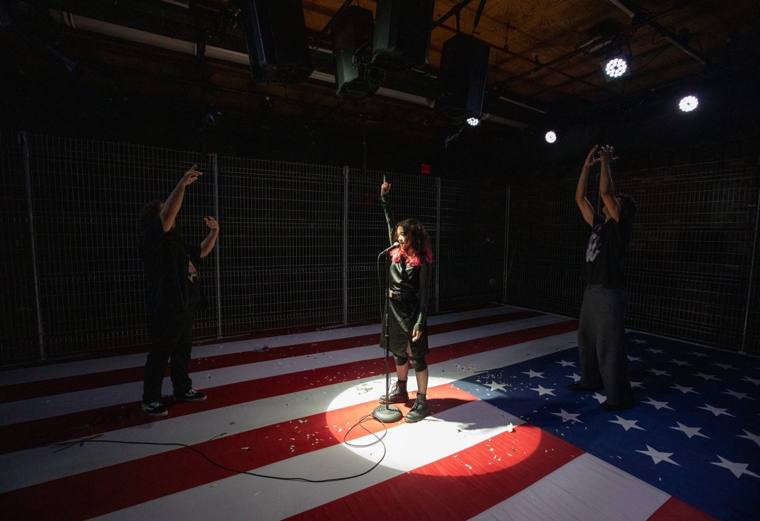 Family: a performer sings into a mic in a spotlight on a stage painted as an American flag, inside a chain-link enclosure, as two others raise their arms