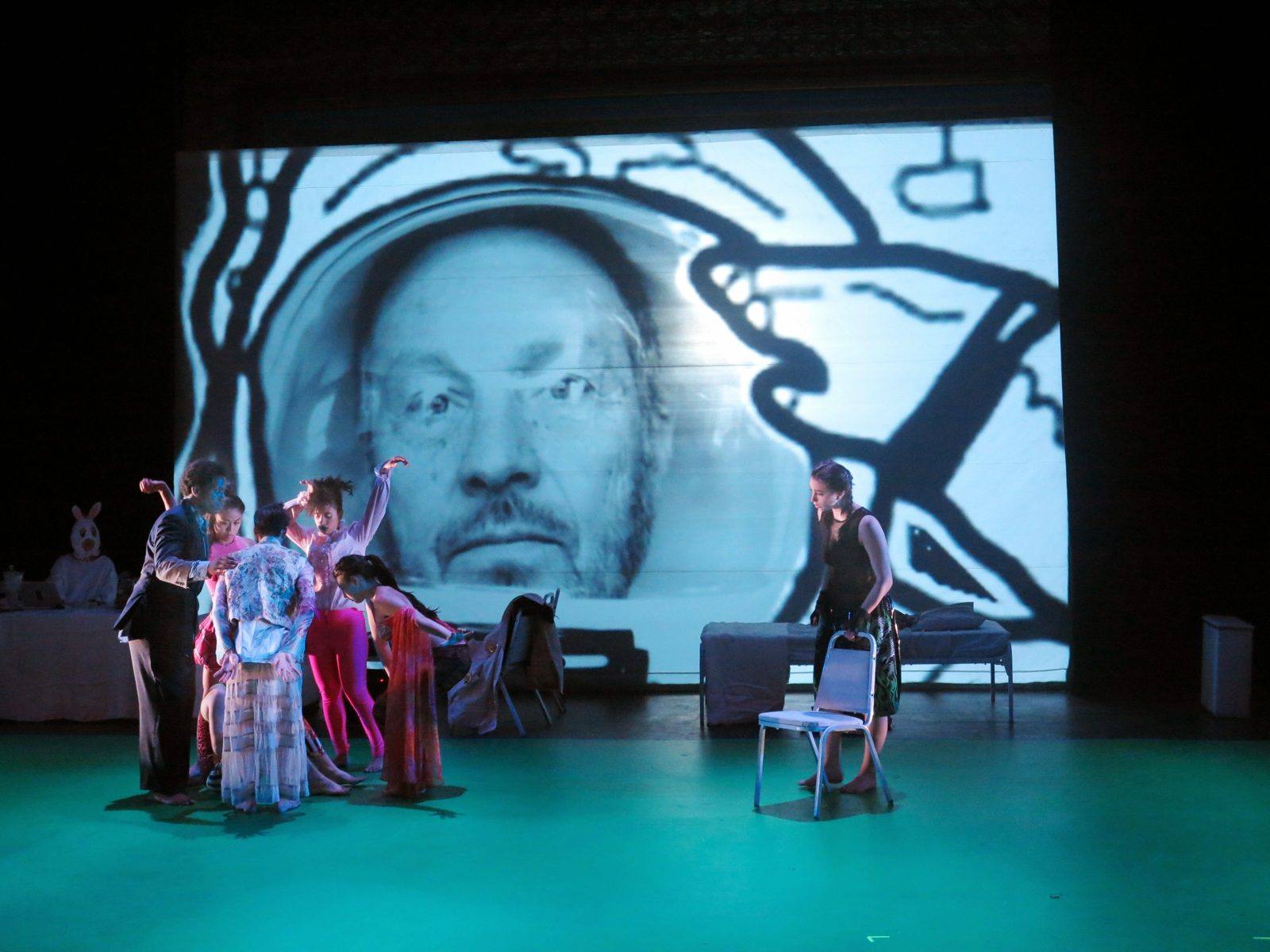 Daily Life Everlasting: dancers on a green floor beneath a giant projected face