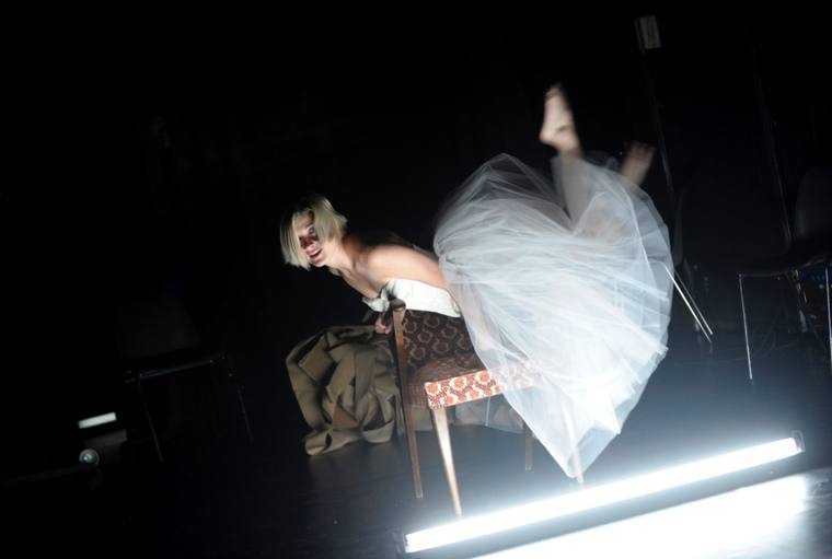 A woman in a tulle skirt tipped over a chair, lit by a fluorescent tube