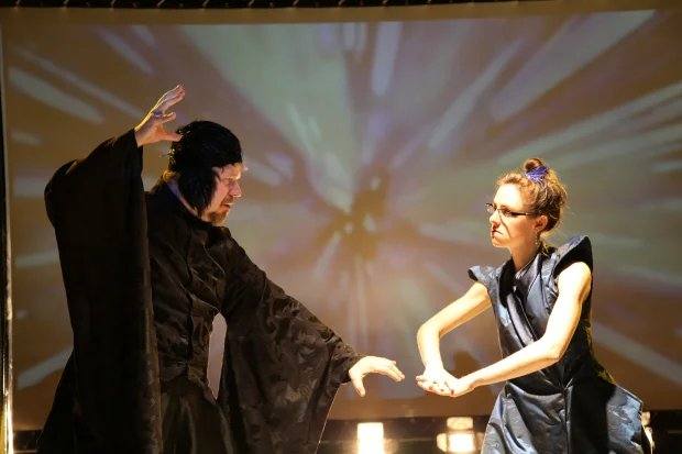 A wizard in black robes and a woman in a metallic vest square off before a starburst projection