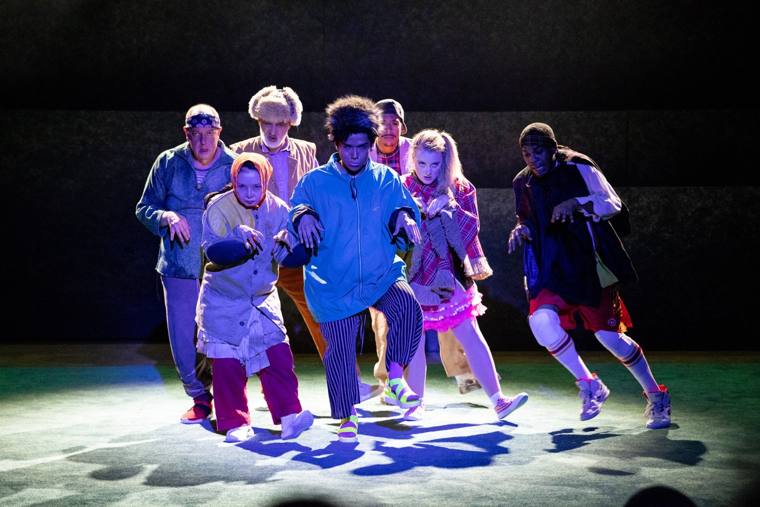 Dom Juan: seven performers in mismatched thrift-store costumes creep forward together in a crouch under blue light