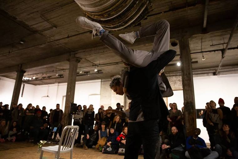 Surveys the Prairie of Your Room: a performer in grey is lifted upside down over Dan Safer’s shoulders in a warehouse ringed by audience