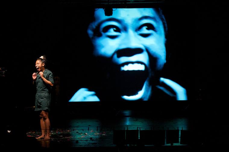 A performer screams beside a giant projection of her own screaming face