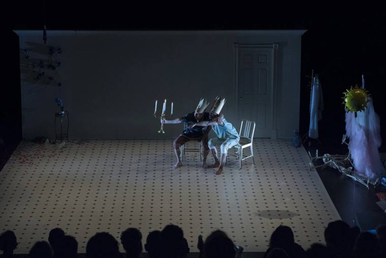 Two performers in paper crowns huddle on chairs holding a candelabra on a white tiled floor