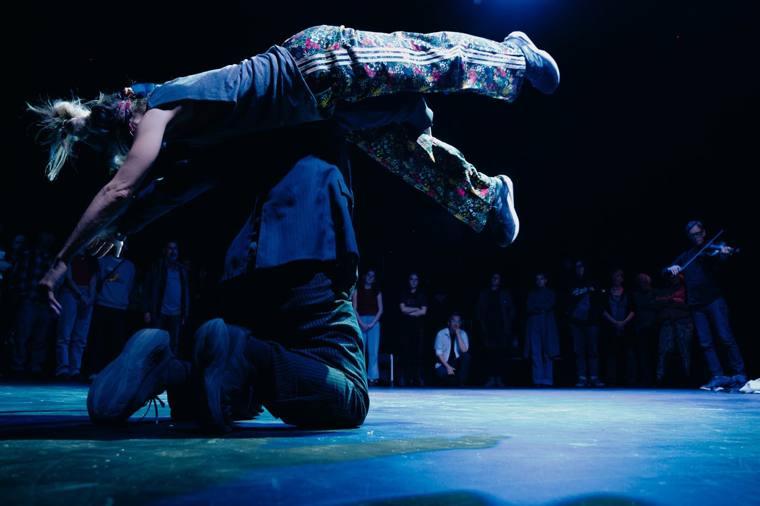 …(And I Feel Fine): a dancer in floral trousers is carried horizontally across another performer’s back, in a blue-lit circle ringed by standing audience