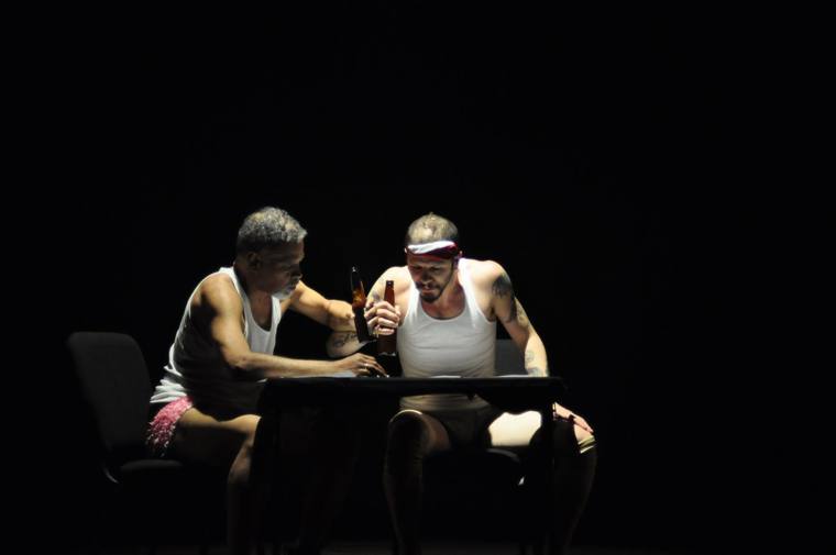 Two men in undershirts share beers at a small table in a pool of light
