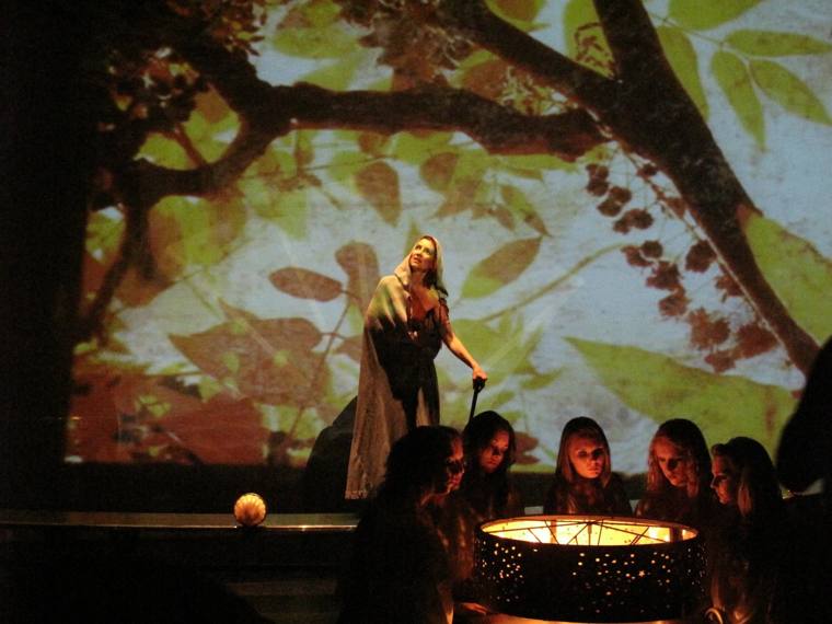 A hooded woman with a cane stands before a projected forest as a chorus gathers around a glowing lamp