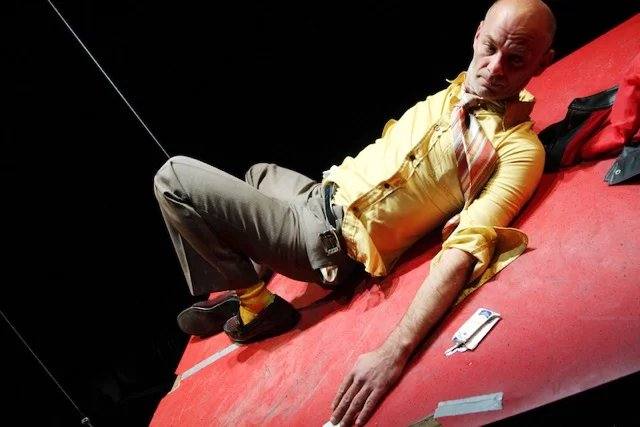 A bald man in a yellow shirt and striped tie sprawls on a red ramp